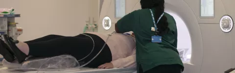 A woman in black trousers and pink top lies down about to have an MRI scan, a radiologists in a green top with long black hair is leaning over her