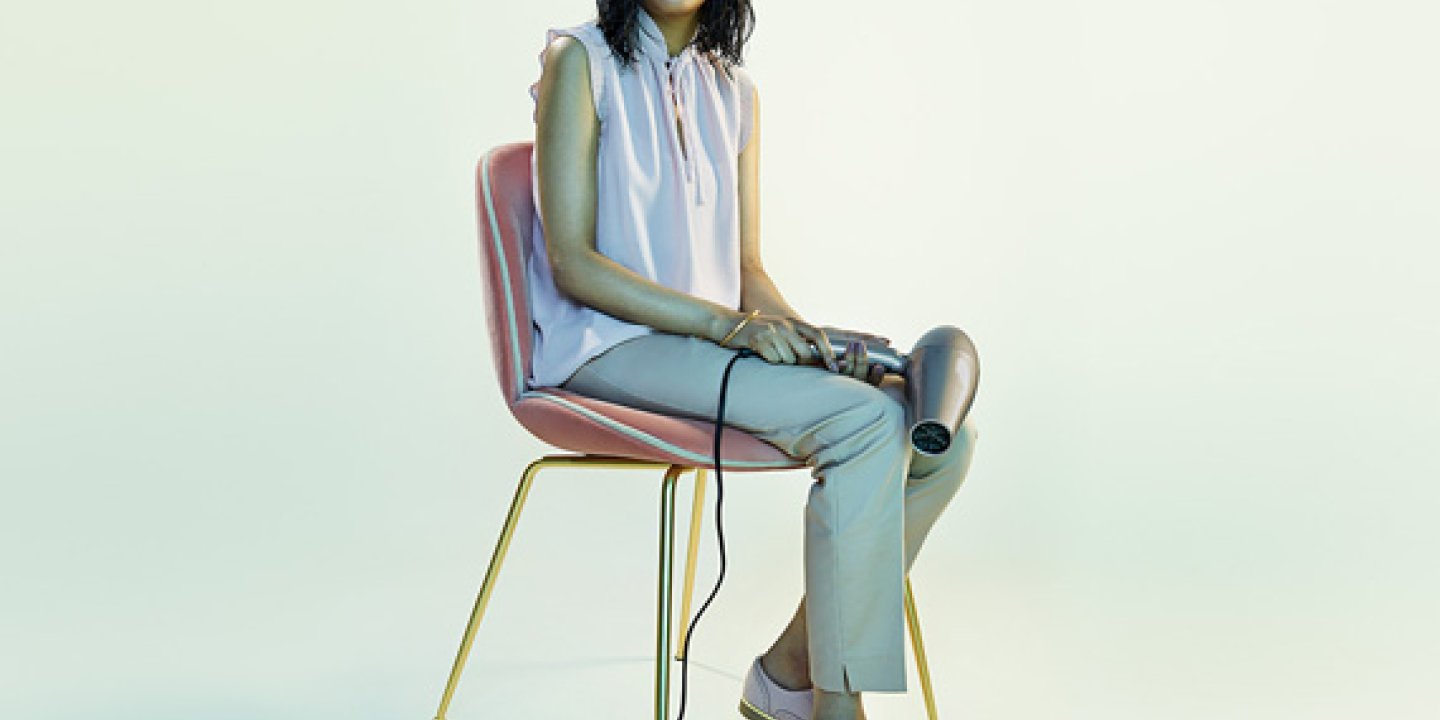 Woman with wet hair sits on a chair holding a hair dryer and looking to camera