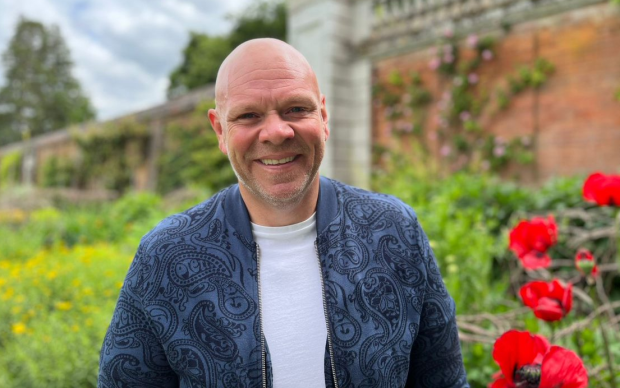 Tom Kerridge stands outside next to some red poppies smiling at the camera