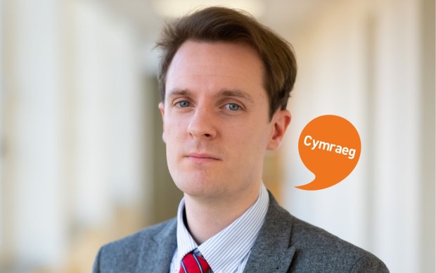 Image shows a man with short brown hair. He is dressed in a shirt, tie and jacket. Cymraeg logo