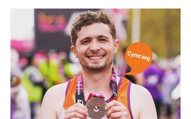 Image shows a man with short brown hair smiling. He is holding a medal after he completed the London Marathon 2023. Cymraeg logo. 