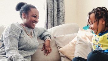 A Black woman living with MS and her son sit together on the sofa, laughing and smiling at each other