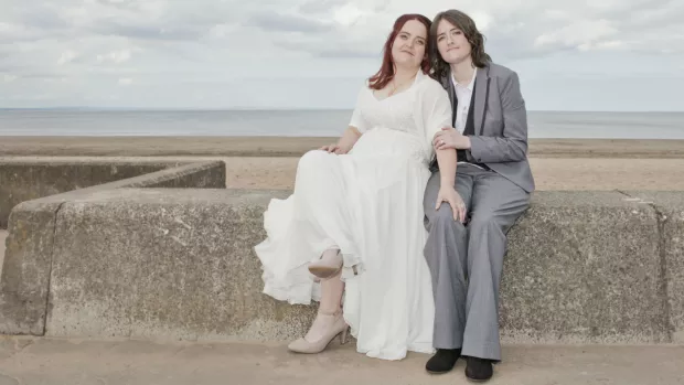 Sarah and Karine on their wedding day, pictured sitting on a wall by the beach. Sarah is wearing a beautiful white wedding dress, Karine is wearing a sharp grey suit.