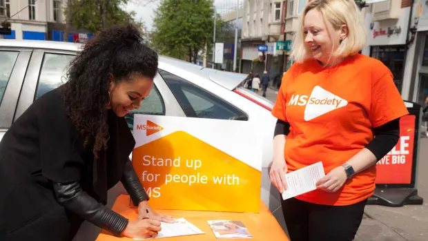 Photo shows a woman signing a petition with another woman in an MS Society t-shirt beside her