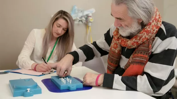 A man with grey hair, wearing a scarf and jumper is sat at a table doing a test involving putting white pegs in holes in a blue tray. A woman with blonde hair wearing a lanyard is taking notes. 