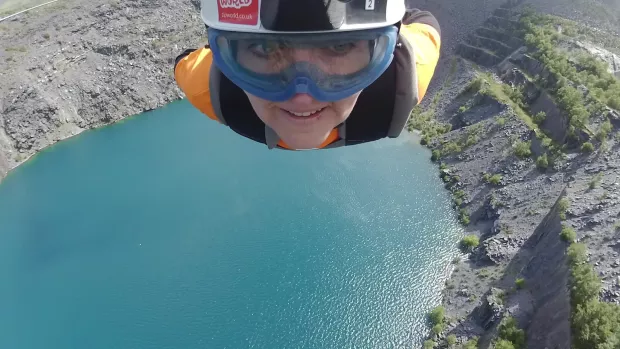 Far below is a water pool in a crater at the top of the image you see just a head wearing goggles and a helmet as the person is suspended by a zip wire