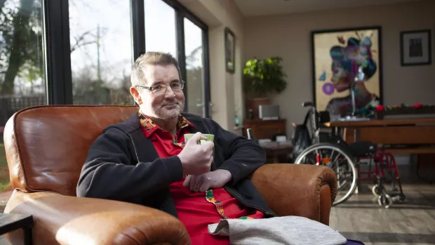 Mark sits in a brown leather armchair holding a mug. Behind him are windowed doors looking into a garden. Mark's wheelchair is in the background. 