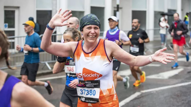 A marathon runner wears an orange MS Society running vest and green bandana. They're smiling and waving with arms spread wide to the camera. 