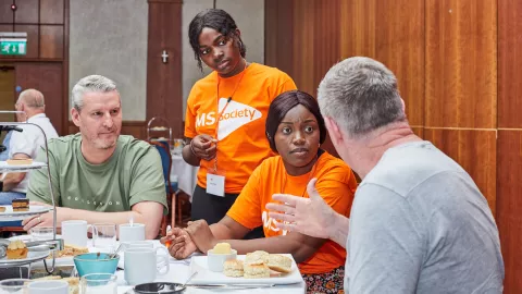 Four people round a table set with an afternoon tea, two of them wear orange MS Society t-shirts, they listen attentively as one of the others talks